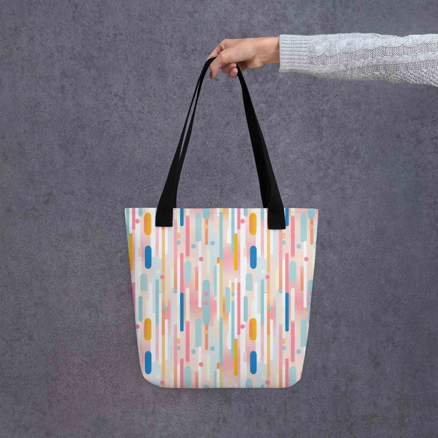 A 15x15 inch tote bag with black 
handles, which design is abstract vertical lines of pastel pink and other pastel shades, held by a lady's hand. Grey background.