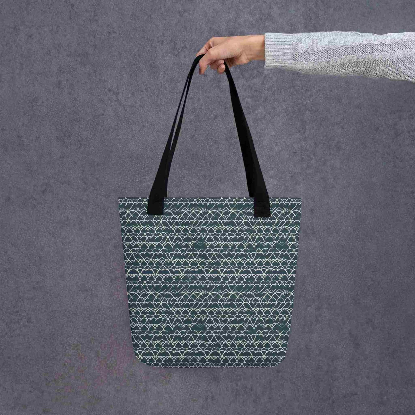 A 15x15 inch tote bag with black handles, which features white geometric lace design that dances across a blue background, held by a lady's hand against a grey wall behind..