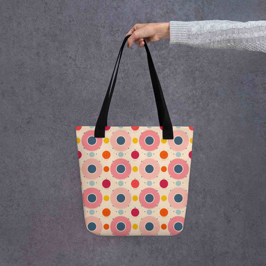 A 15x15 inch tote bag with blac handles, which features an artsy design of pastel candy pops of color in a beige background, held by a lady's hand. Grey background.