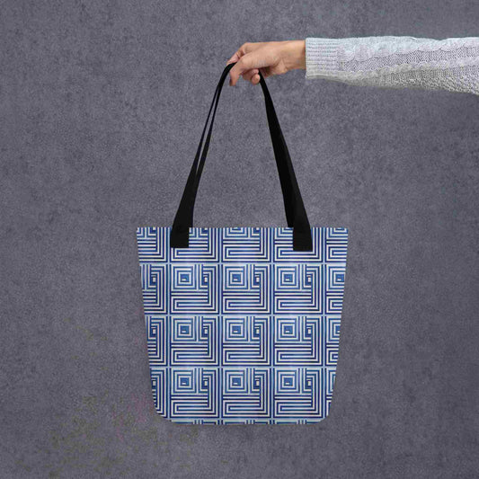 A 15x15 inch tote bag with black handles, which features a blue op art illusion on a white background, held in a lady's hand. Grey background.