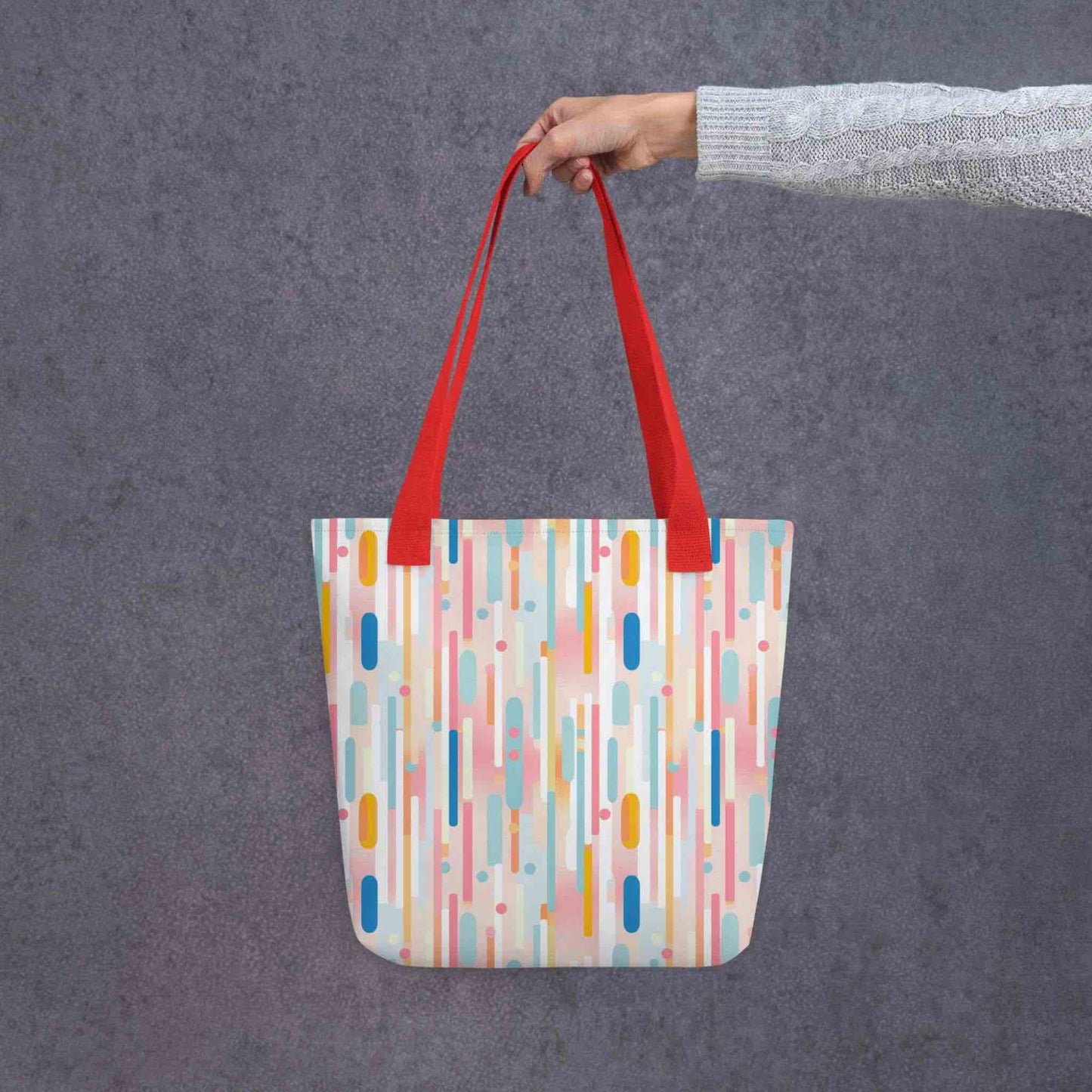 A 15x15 inch tote bag with red handles, which design is abstract vertical lines of pastel pink and other pastel shades, held by a lady's hand. Grey background.