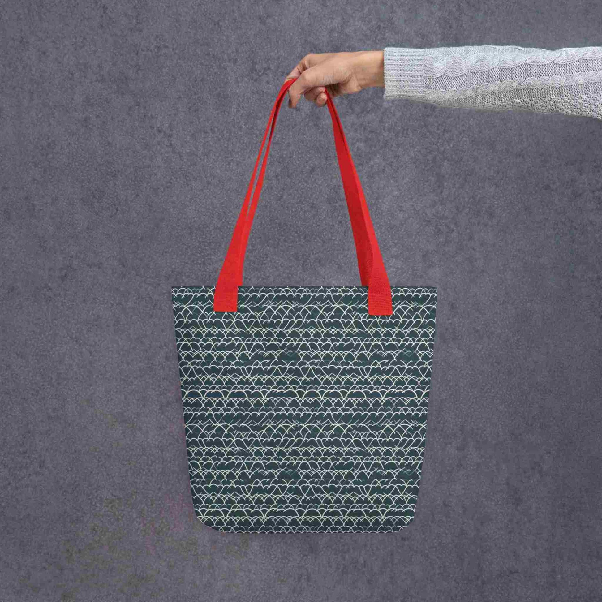 A 15x15 inch tote bag with red handles, which features white geometric lace design that dances across a blue background, held by a lady's hand against a grey wall behind..