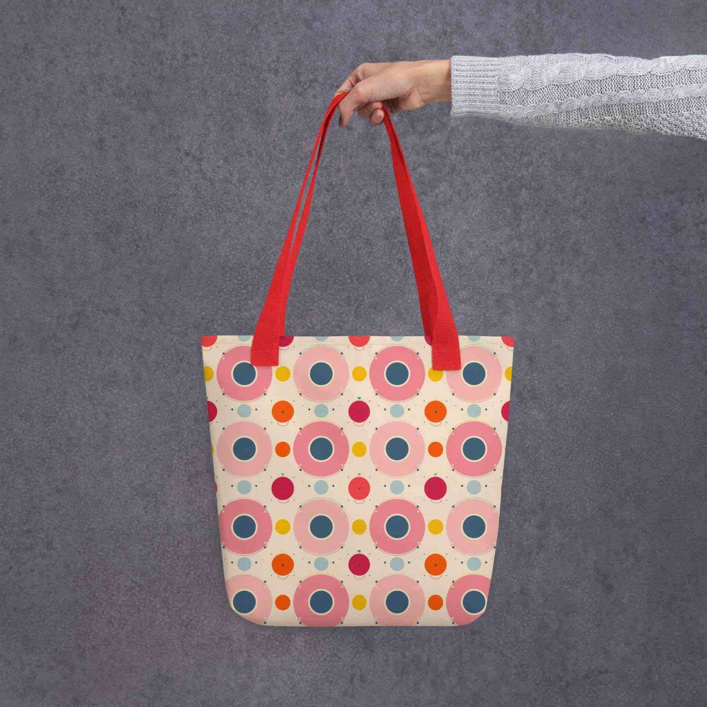 A 15x15 inch tote bag with red handles, which features an artsy design of pastel candy pops of color in a beige background, held by a lady's hand. Grey background.