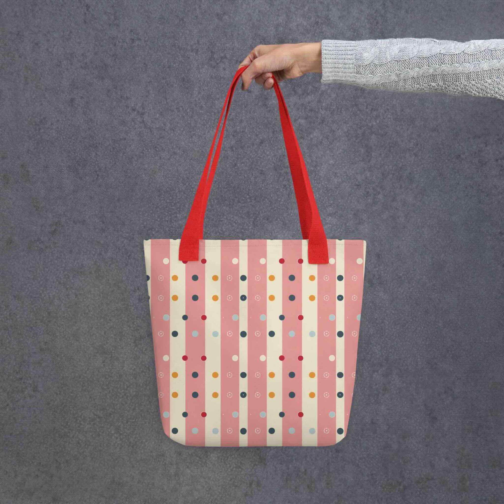 Pink striped tote bag with red handles, which features pink and beige stripes with colourful pokka dots, held by a lady's hand. Dimension 15x15 inches. Grey background.