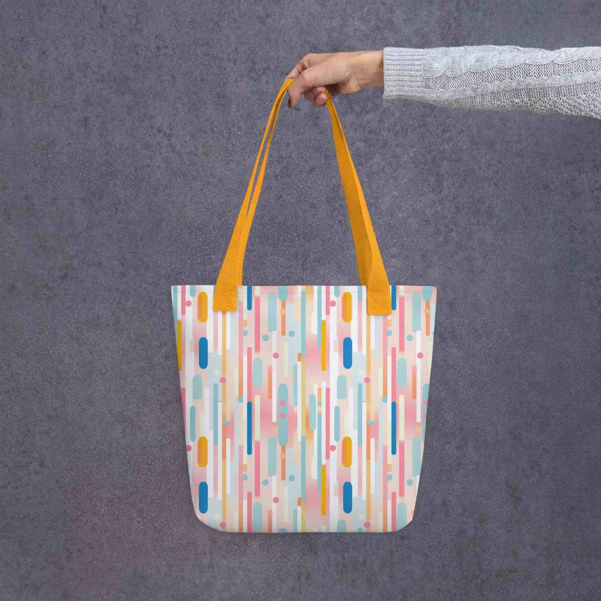 A 15x15 inch tote bag with yellow handles, which design is abstract vertical lines of pastel pink and other pastel shades, held by a lady's hand. Grey background.