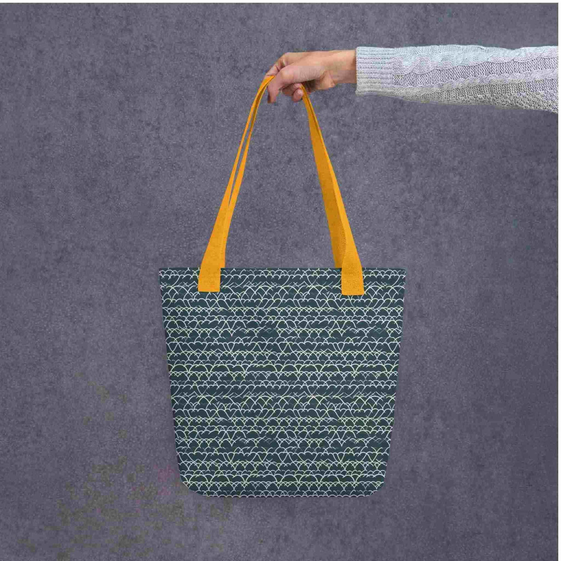 A 15x15 inch tote bag with yellow handles, which features white geometric lace design that dances across a blue background, held by a lady's hand against a grey wall behind..