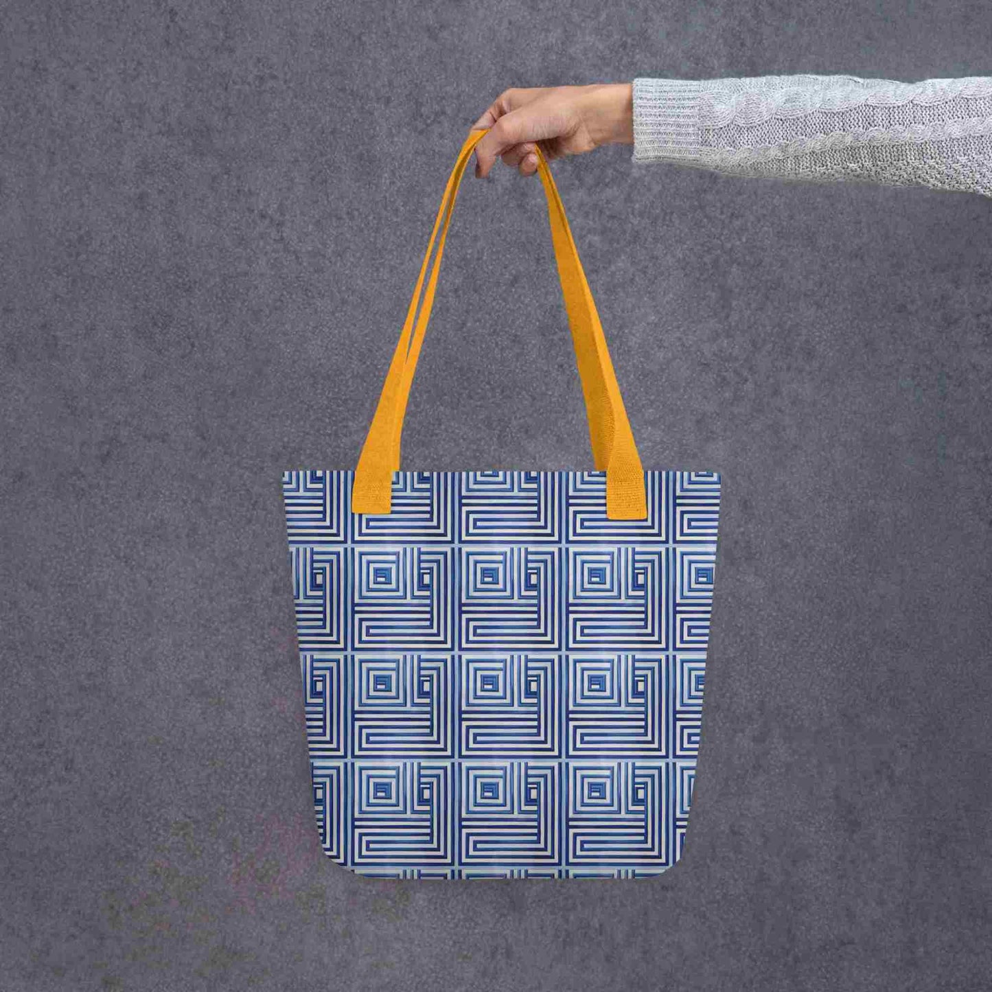 A 15x15 inch tote bag with yellow handles, which features a blue op art illusion on a white background, held in a lady's hand. Grey background.