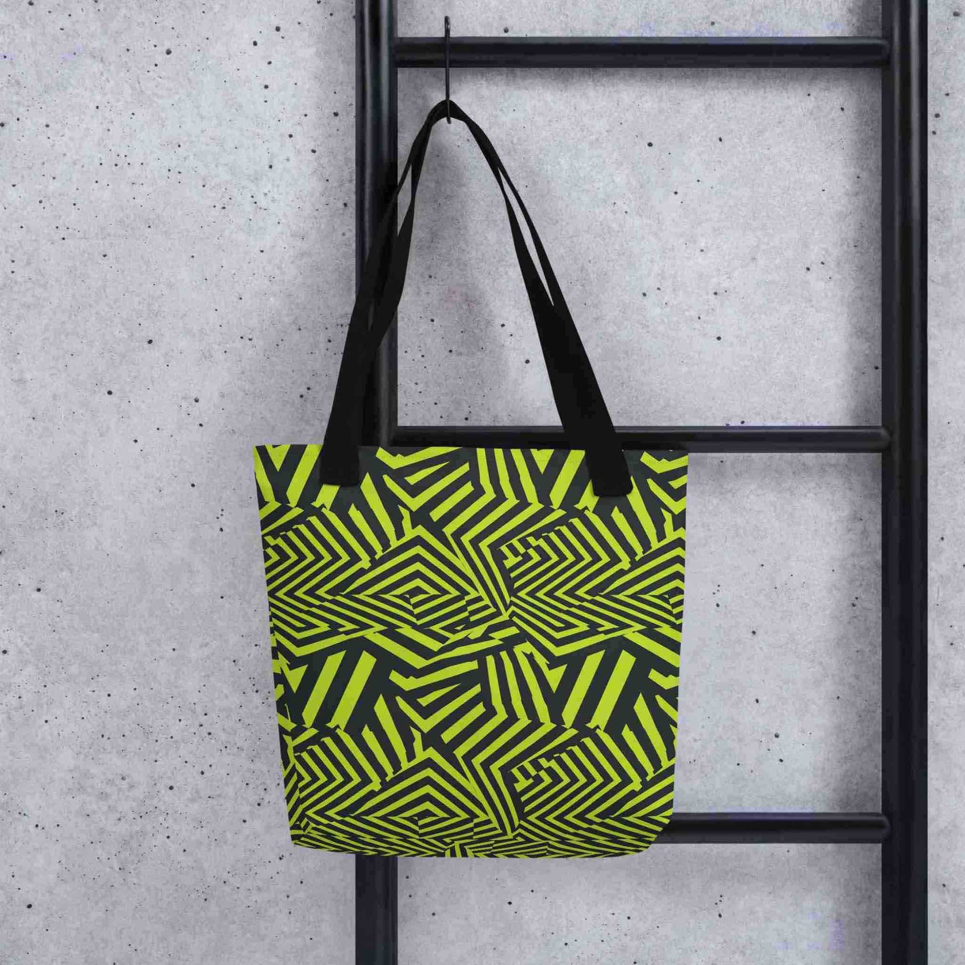 An avant garde tote bag with black handles, which features bold black and green op art illusion, hooked onto a black ladder. Size is 15x15 inch. Grey background.