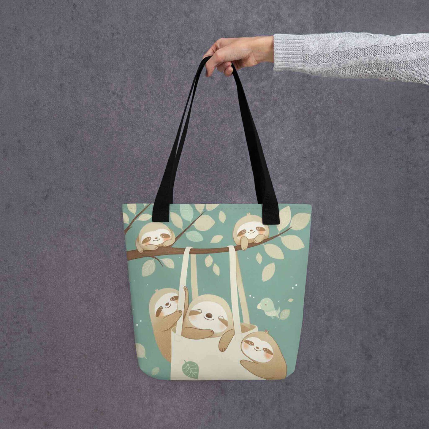 The cutest sloth tote bag you'll ever carry! This charming, sophisticated design captures the tranquility of the forest, with black shoulder handles, held in a hand. Grey background.