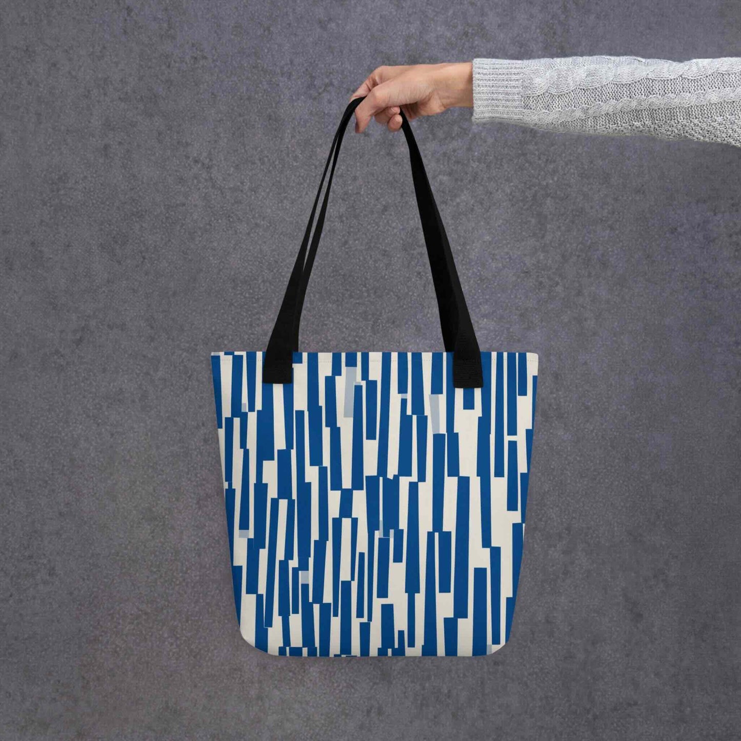 The tote bag blue, where rhythm meets charm. This abstract design features a chaotic yet charming cascade of navy shapes, resembling musical staccato notes played against a soft cream backdrop. The bag is held i a hand against a grey background.
