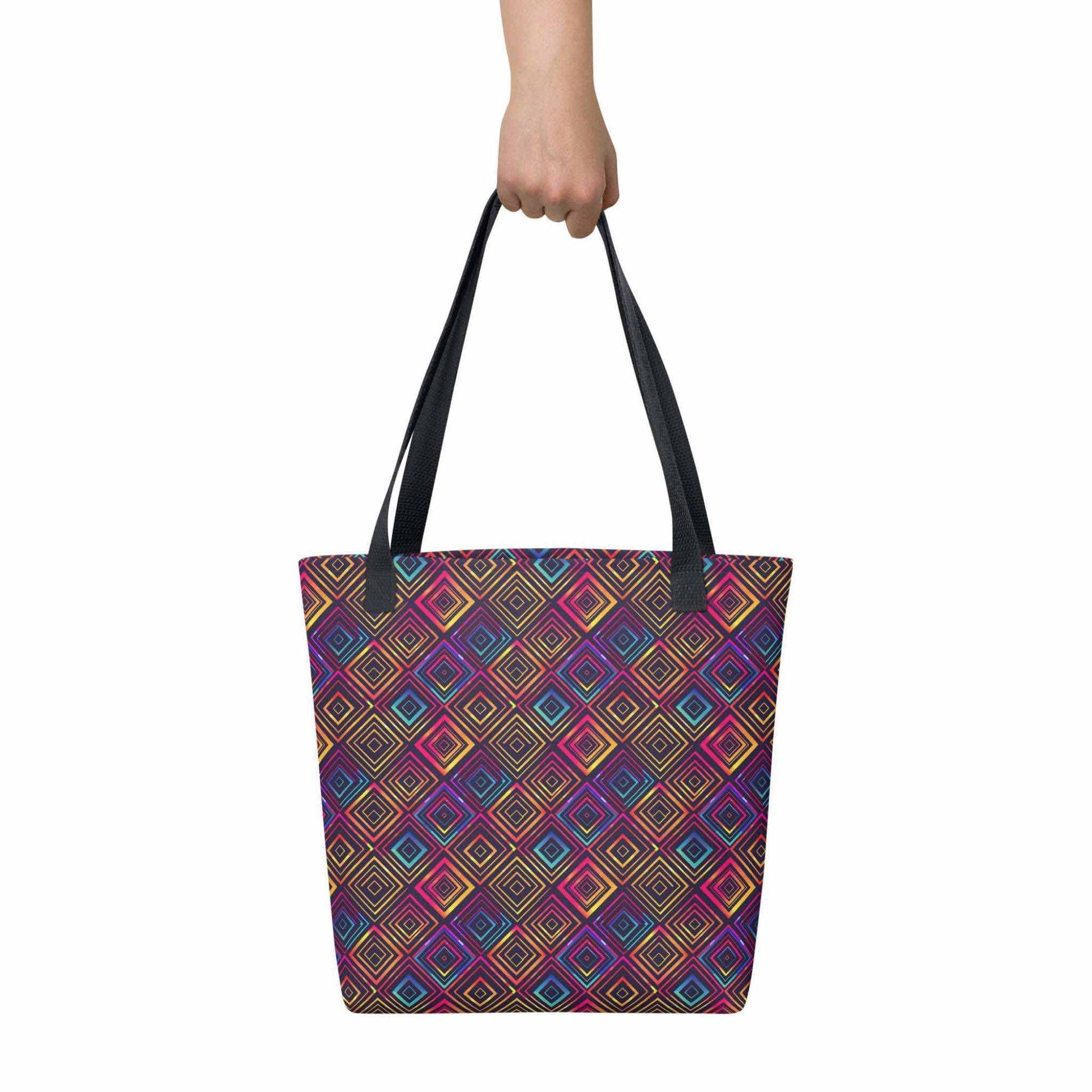 Fashion tote bag with black handles, which features a neon tapestry design in a sophisticated blend of colors, held in a hand. Dimension 15x15 inches. White background.