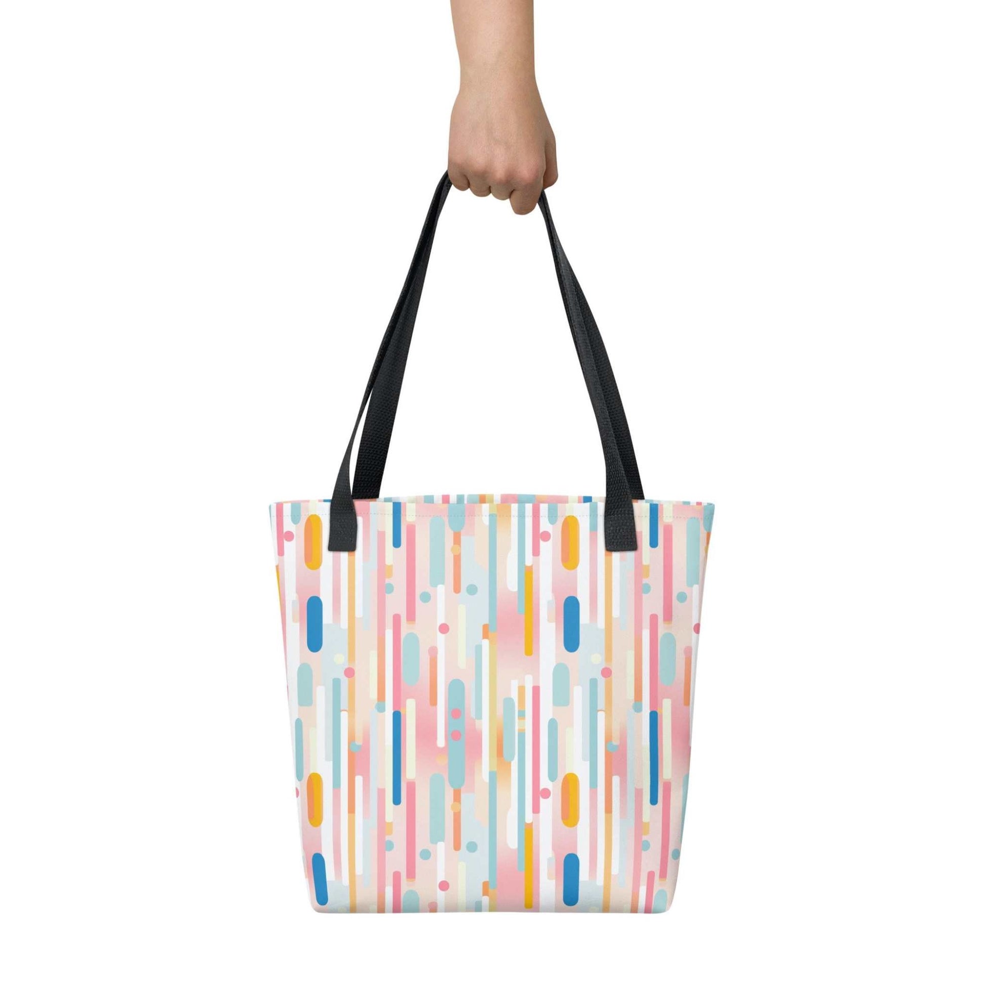 A 15x15 inch tote bag with black handles, which design is abstract vertical lines of pastel pink, and other pastel colours, held by a lady's hand. White background.